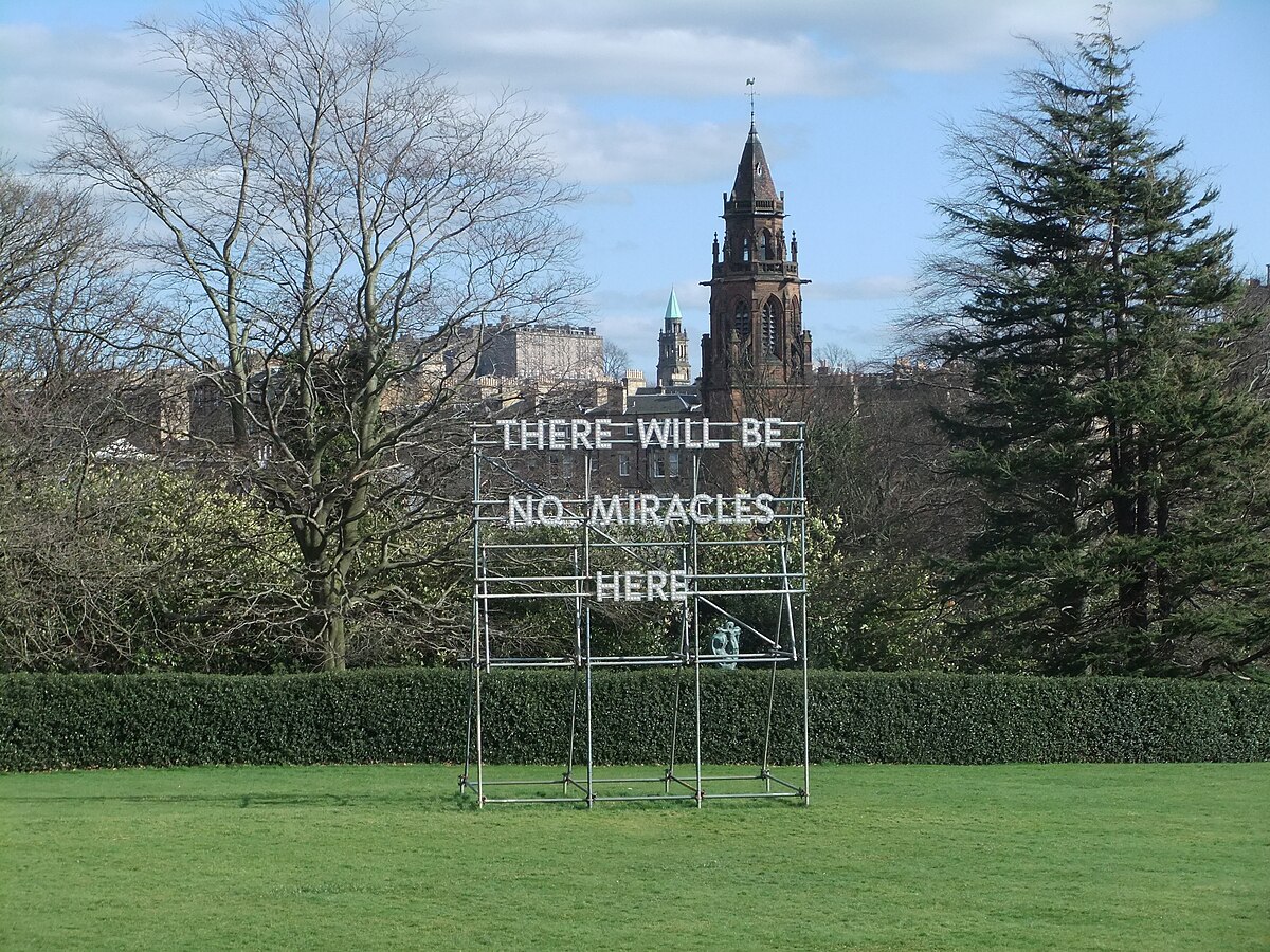 Neon metal sign in a grassy area reads “THERE WILL BE NO MIRACLES HERE,” with trees and a church steeple in the background.