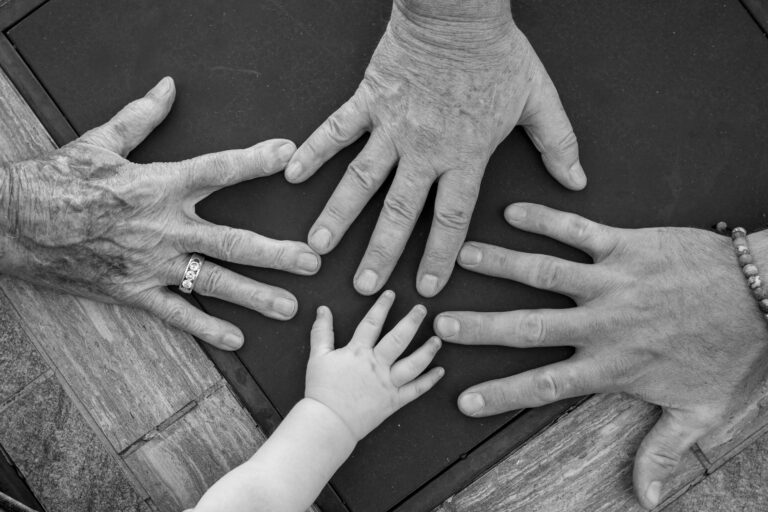 3 adult hands and 1 child hand with palms down on a table, arranged in group.